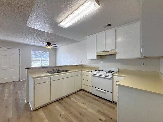 A renovated kitchen with white cabinets, a white gas range, a double sink under the window and light wood-look flooring